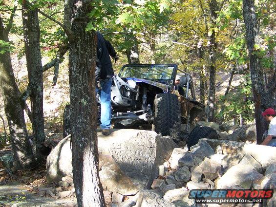 Off-road vehicle with roll cage navigating rocky terrain, person standing nearby