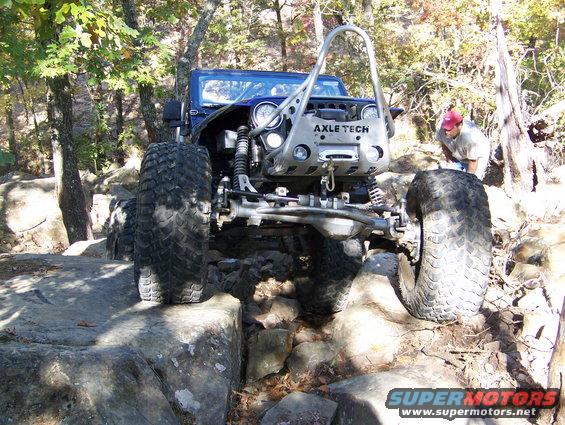 Blue Jeep Wrangler with large tires and custom front bumper on rocky terrain. 'AXLE TECH' is visible on the bumper