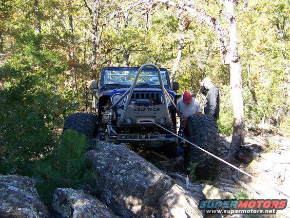 Blue Jeep Wrangler with large tires navigating rocky terrain, two people nearby
