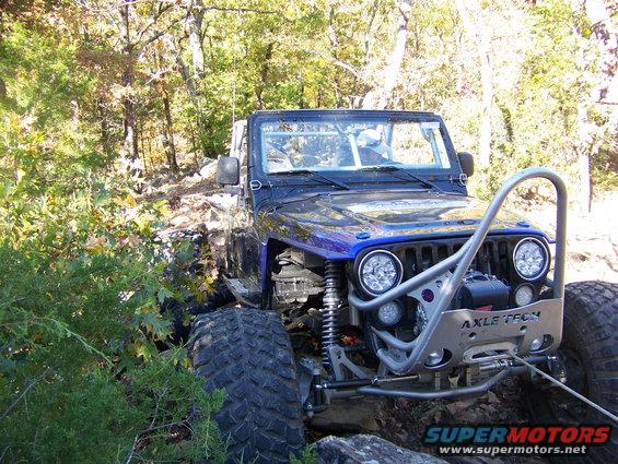 Modified blue Jeep Wrangler with large tires and custom bumper, winching on a rocky trail