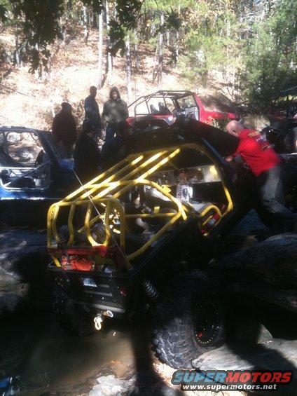 Black off-road vehicle with yellow roll cage, tilted on rocks in water. A man in a red shirt pushes it