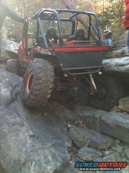 Red off-road vehicle with roll cage navigating rocky terrain; another vehicle visible in background