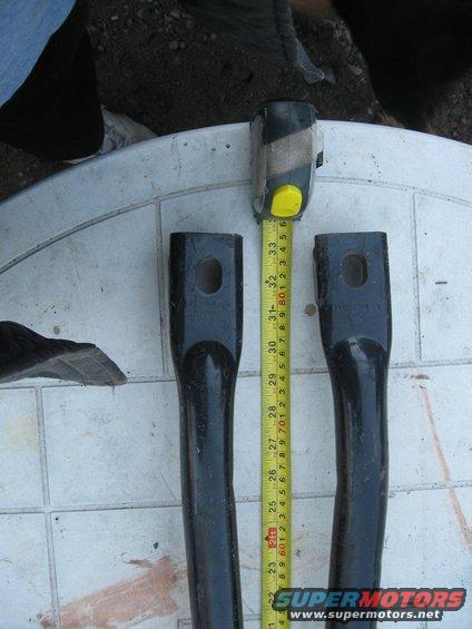 Two black metal parts with holes, measured by a yellow tape measure