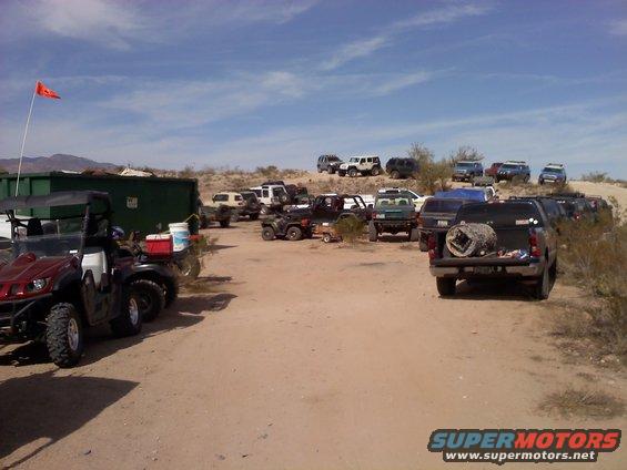 Desert scene with many parked SUVs, trucks, and ATVs under a blue sky with scattered clouds