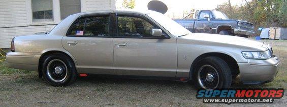 Tan 2000 Mercury Grand Marquis sedan, side view, black wheels, parked on gravel