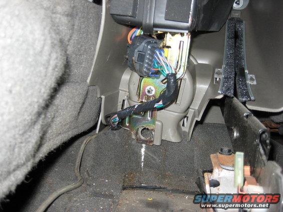 Wiring harness connected to a metal bracket inside a vehicle's interior, with gray carpet and plastic trim visible