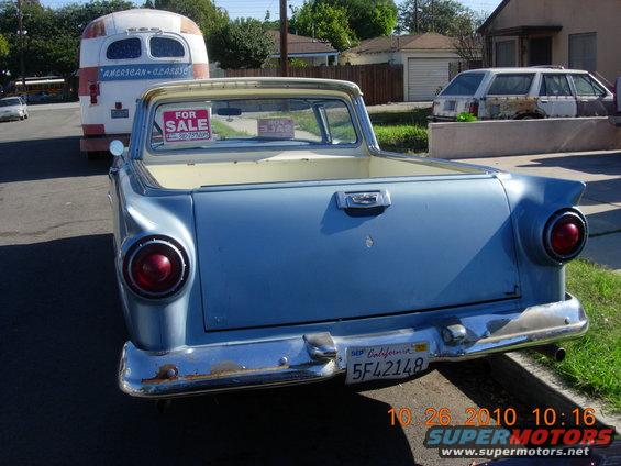 Rear view of a blue 1957 Ford Ranchero pickup with 'FOR SALE' signs in the window. A vintage bus is parked behind it