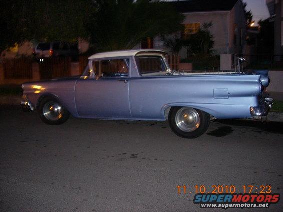 Light blue 1957 Ford Ranchero parked on a street at night. It has a white roof and bed cover