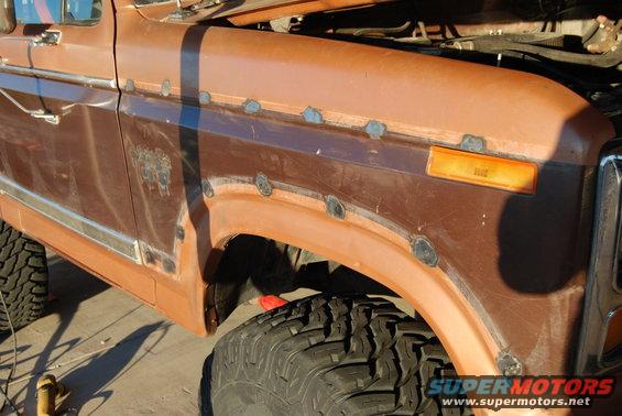 Side view of a brown Ford truck fender with rust spots and new welds. Hood is open, showing engine parts