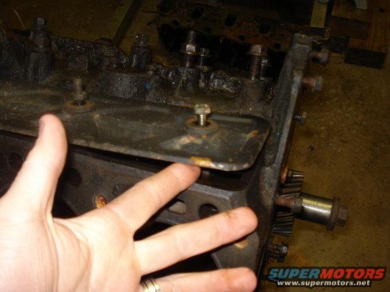 Close-up of an engine block with gears visible, a hand is pointing to a metal plate