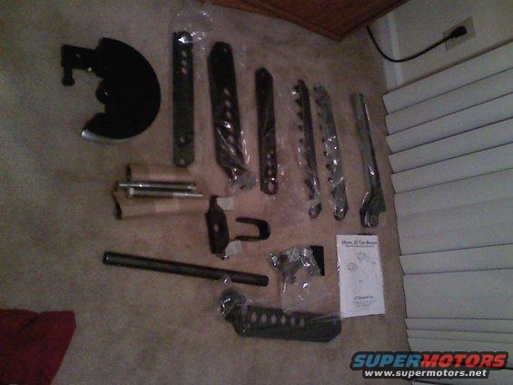 Various metal vehicle parts laid out on carpet, including brackets, a brake rotor shield, and a long metal rod. An instruction manual is visible