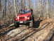 1991 Ford Bronco - A red Jeep Wrangler navigates a challenging off-road path through a forest with bare trees.
