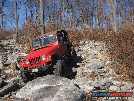 Red Jeep Wrangler on a rocky trail, front-left view, with aftermarket wheels and tires