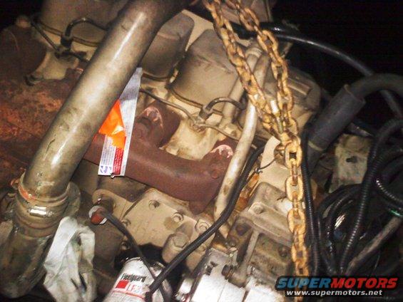 Engine block with exhaust manifold, oil filter, and lifting chains. Orange tag attached to manifold