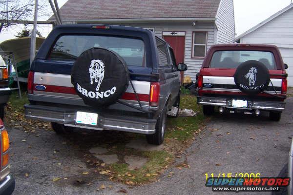 Two Ford Broncos parked in a driveway, rear view. One is blue and silver, the other is red