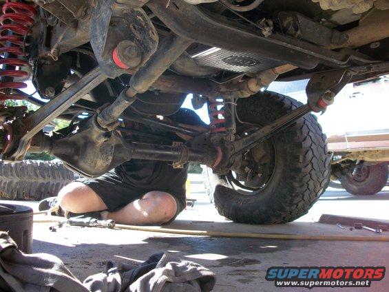 Underbody view of a vehicle with a person working on the suspension and drivetrain. Another vehicle is visible in the background