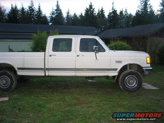 White crew cab pickup truck, side view, parked on grass with trees and buildings in background