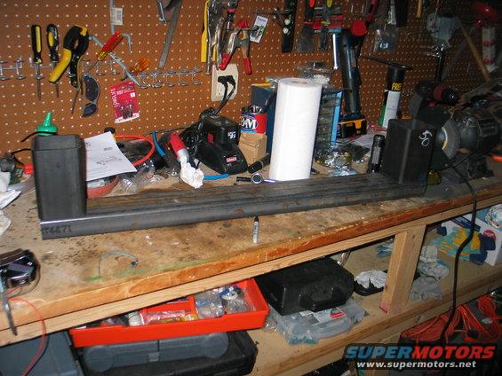 Metal bar with brackets and "T4471" etched on it, sitting on a workbench with tools and supplies