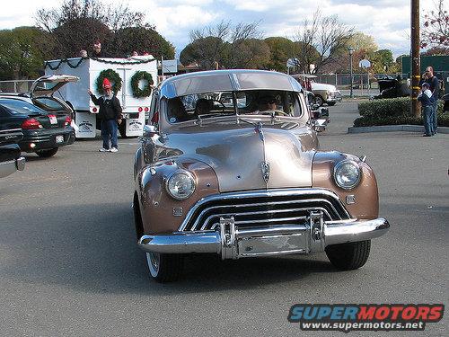 Front view of a two-tone brown and tan vintage car with chrome grille and bumper. Other vehicles and people are visible in the background