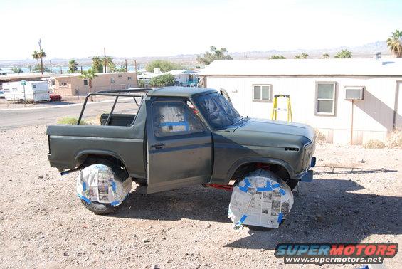 1980 Ford Bronco with doors open, wheels taped off for painting. Ladder and trailer in background