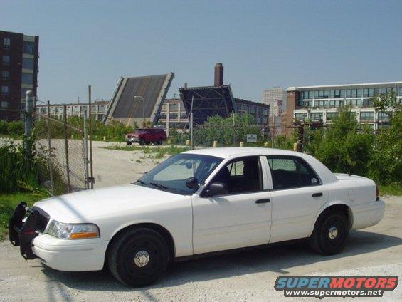 2003 Ford Crown Victoria police car parked on dirt lot with industrial buildings and a drawbridge in background