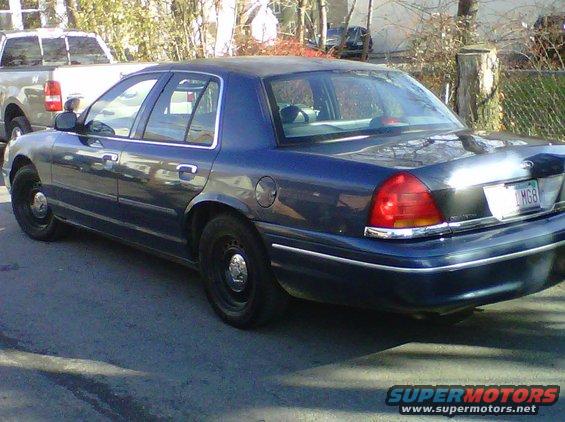 Dark blue Ford Crown Victoria sedan, police interceptor model, parked on street. Another pickup truck is visible behind it