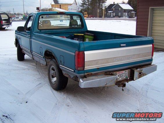 Teal pickup truck with silver tailgate trim and a dented chrome bumper, seen from the rear