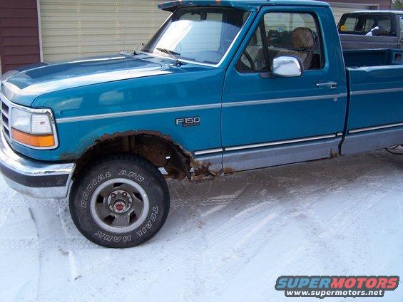 Teal 1994 Ford F-150 pickup truck with rust on the fender and rocker panel. Another truck is visible in the background