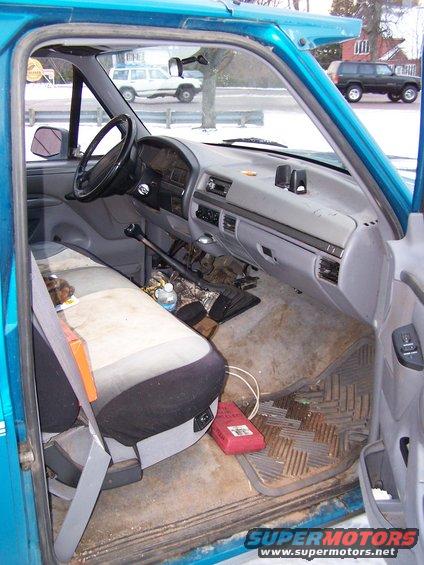 Interior of a 1994 Ford F-150 pickup truck showing the driver's seat, dashboard, and steering wheel. Another white SUV is visible through the windshield