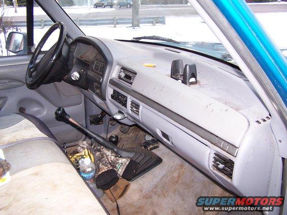 Interior of a truck cab, showing the dashboard, steering wheel, and gear shifter. There are boots and camouflage clothing on the floor