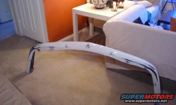 White car bumper with black trim and some markings on it, sitting on the floor indoors