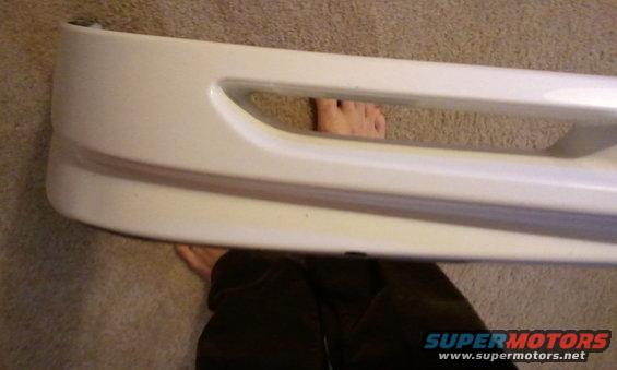 White car bumper with a horizontal opening, seen from the side. Feet are visible through the opening