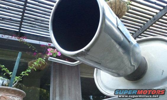 Close-up of a chrome exhaust pipe and muffler, with a potted plant and wooden pergola in the background. Supermotors.net logo visible