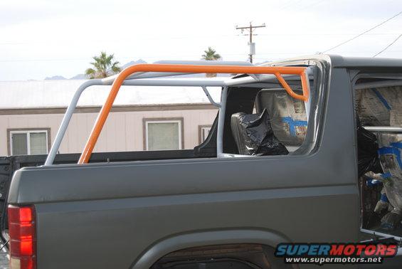 Gray Ford Bronco with orange and white roll cage, rear side view. Interior is covered with plastic and tape