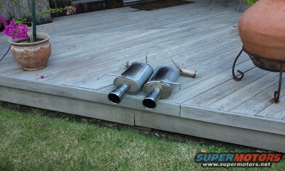 two chrome exhaust mufflers with bent tips resting on a wooden deck next to a potted plant and a clay chiminea