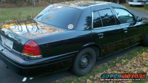 Black sedan, rear quarter view, parked on grass with fallen leaves. License plate visible