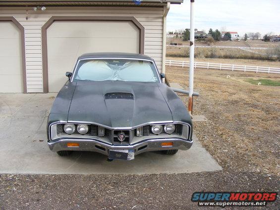 Dark green coupe with a hood scoop and chrome grille, parked in front of a garage