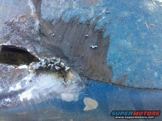 Close-up of a blue metal panel with rust and welding marks, showing a hole and rough welds