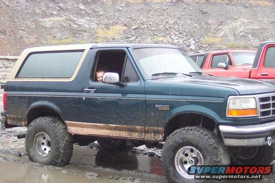 Green Ford Bronco with a child in the driver's seat, parked in mud. Red truck visible behind it