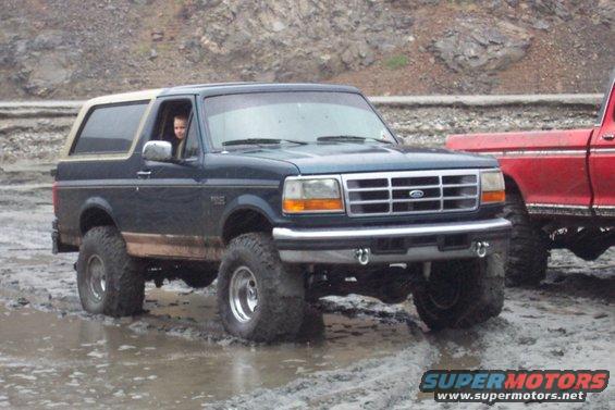 Dark blue 1995 Ford Bronco with a tan top, lifted suspension, and large tires driving through mud. A red pickup truck is partially visible to the right
