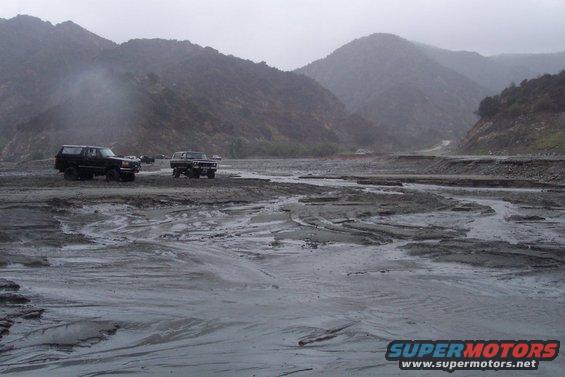 Two dark SUVs navigate a muddy, rocky riverbed with mountains in the background