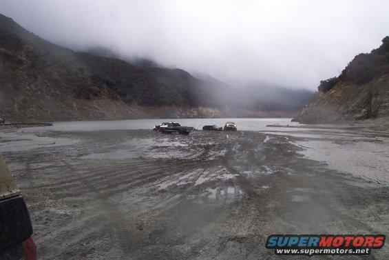 Several vehicles stuck in mud near a lake on a foggy day