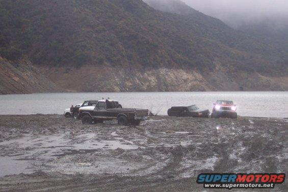 Several vehicles stuck in mud near a body of water with a hill in the background