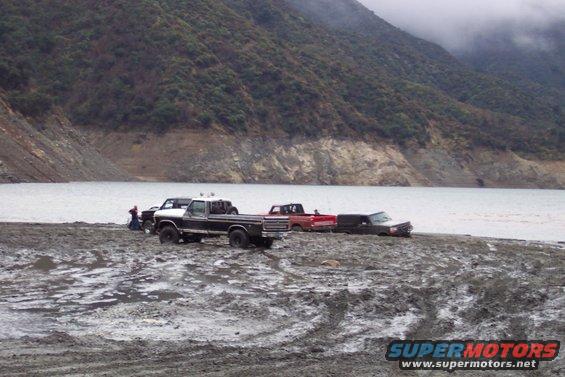 Several pickup trucks are stuck in mud near a body of water with a hillside in the background