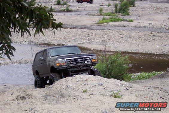 1995 Ford Bronco climbing a dirt mound in a rocky stream bed. Another vehicle is visible in the background