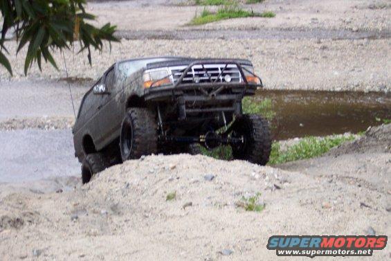 Off-road SUV with large tires climbing a dirt hill next to water. Custom front bumper and roll cage visible