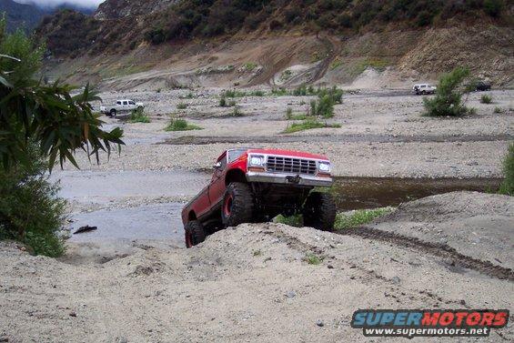 Red lifted pickup truck with large tires driving through a rocky stream bed. Other vehicles are visible in the background