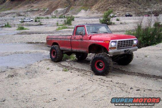 Red pickup truck with huge tires on dirt road. Another white truck in background