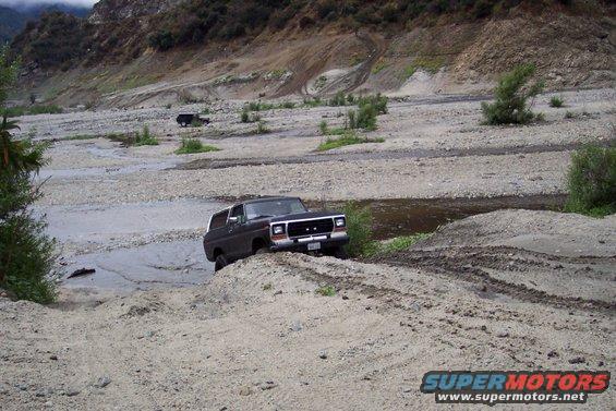 Dark SUV on dirt mound near water, with another SUV in background
