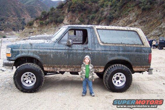 Mud-covered 1995 Ford Bronco parked off-road with a child standing next to it. Another vehicle is visible in the background